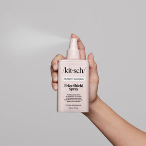 KITSCH Humidity Blocking Frizz Shield Spray — hand holding bottle from below with nozzle up, spray mist visible — product action shot on light gray background — Humidity-Blocking | Frizz Shield Spray — Formulated with GLASSPLEX™ — For sleek, shiny blowouts — 6 fl. oz. / 177mL — Hydrates without weighing hair down | Protects hair from heat up to 266°F/130°C