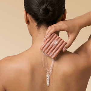 Woman lathering Kitsch Amber Shores Body Wash Bar on shoulder with sudsy foam on a beige background