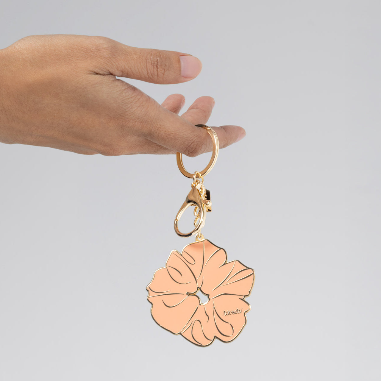 Gold metal enamel keychain depicting the kitsch Terracotta Scrunchie with gold outline, design, and hardware held by a hand against a gray background