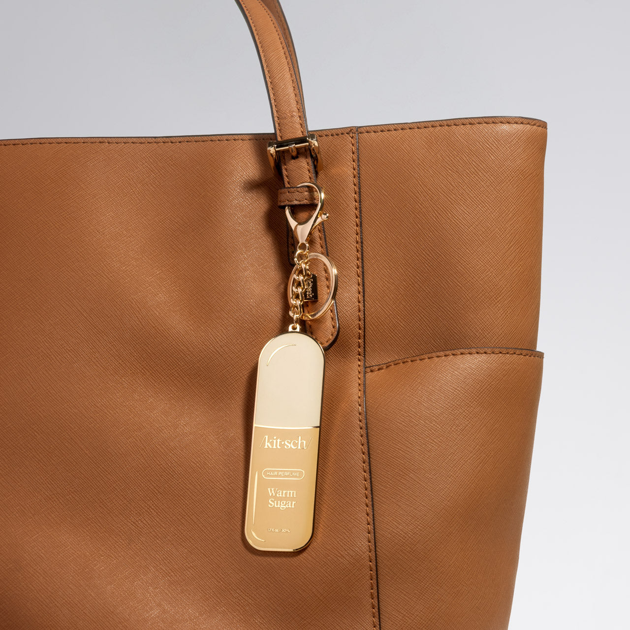 Brown leather handbag with a metal enamel keychain depicting the kitsch Warm Sugar hair perfume with gold outline, design, and hardware attached on a light gray background