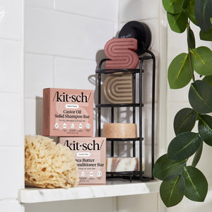 KITSCH Shea Butter Hydrating Solid Conditioner Bar — lifestyle bathroom shot with bar and box on black metal shower caddy — styled with KITSCH Castor Oil Solid Shampoo Bar, natural sea sponge, and tropical plant — white tile background — Richly hydrates dry, thirsty strands — PH balanced and safe for color-treated hair — Reduces single-use plastic