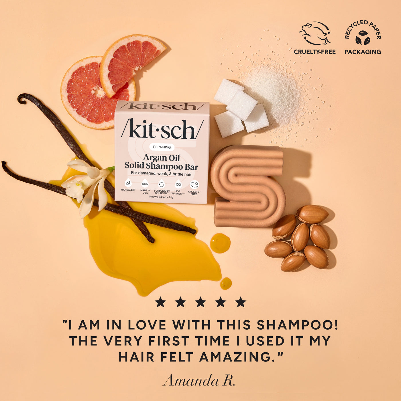KITSCH Repairing Argan Oil Shampoo Bar — ingredient lifestyle flat lay on warm peach background — bar and retail box with grapefruit, vanilla beans, argan nuts, and white sugar crystals — Cruelty-Free | Recycled Paper Packaging — 5-star customer review: "I AM IN LOVE WITH THIS SHAMPOO! THE VERY FIRST TIME I USED IT MY HAIR FELT AMAZING." — Amanda R. — Packed with antioxidant-rich argan oil to boost moisture without weighing it down